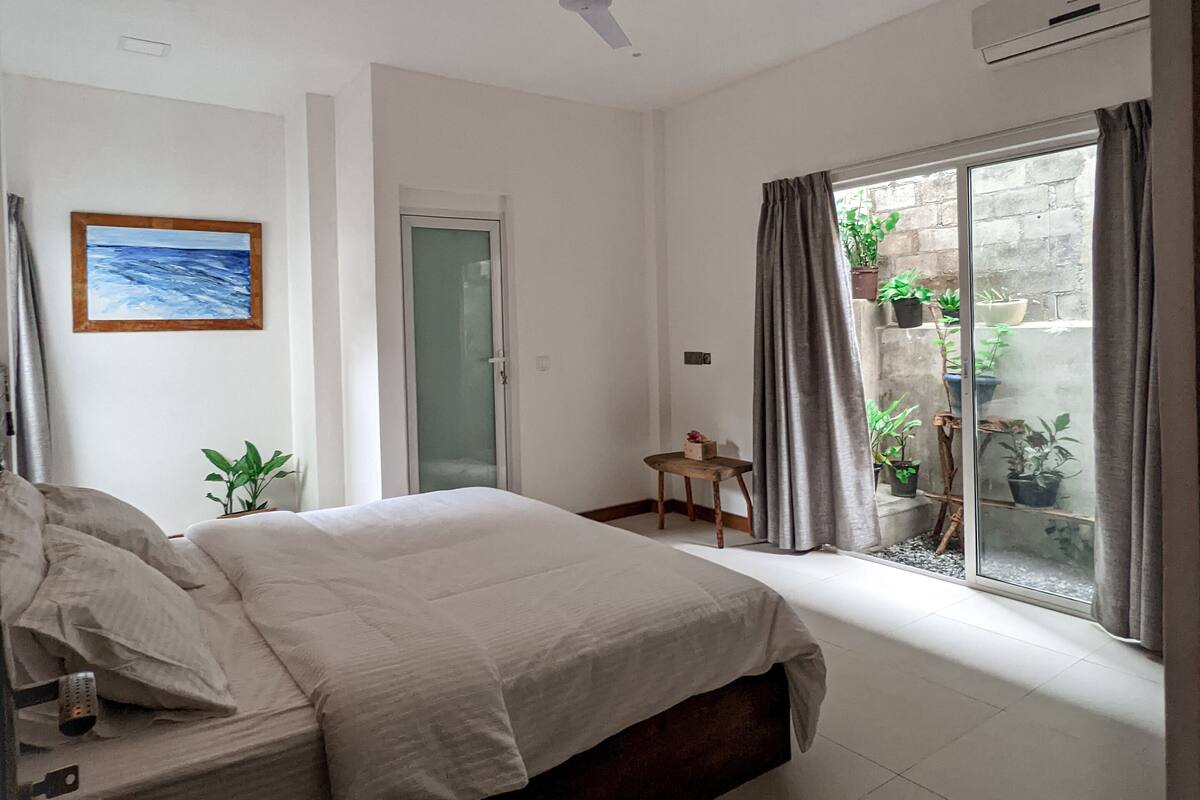 Un dormitorio con cama, ventilador de techo, ventana con cortinas, una puerta y una mesita con una planta.