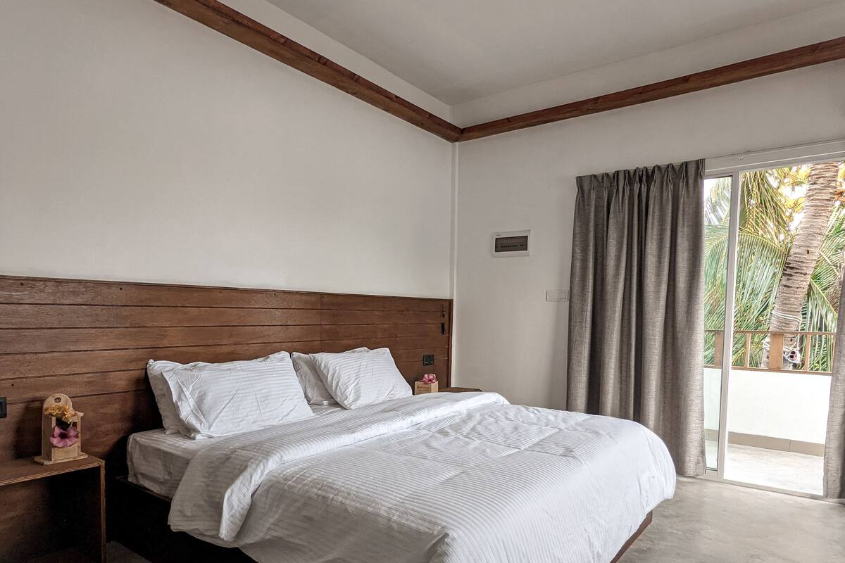 Un dormitorio con cabecera de madera, cama con ropa de cama blanca, mesita de noche, ventilador de techo y vista a través de una puerta corrediza de vidrio.