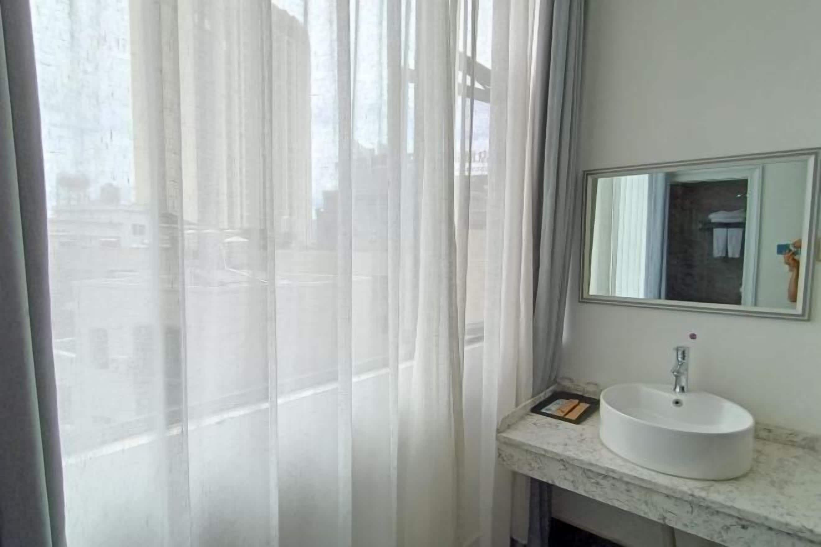 A bathroom with a white sink, a mirror, and a window with sheer curtains.