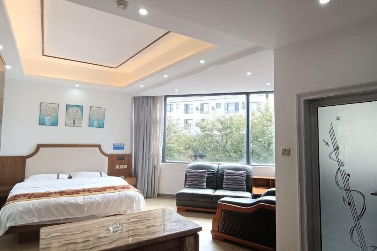 Quarto de hotel moderno com uma cama grande, um sofá, uma mesinha de mármore e uma janela com vista para edifícios e árvores.