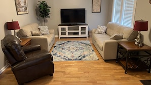 Ein Wohnzimmer mit braunen Ledersesseln, beigen Couches, Holzboden, Fernseher und einer Pflanze.
