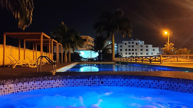 A nighttime swimming pool with clear water, surrounded by palm trees and a building with illuminated windows.