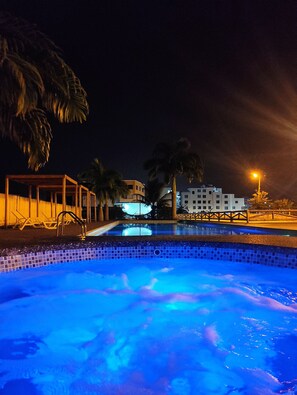 A nighttime swimming pool with clear water, surrounded by palm trees and a building with illuminated windows.