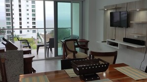 A modern living room with a wooden dining table, chairs, a TV mounted on a wooden panel wall, and a large sliding glass door leading to a balcony with a view of the ocean and buildings.