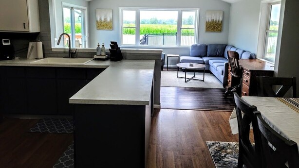 A modern kitchen with a large island, a dining area with a sofa and table, and a view of a green field through large windows.
