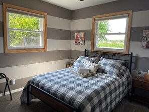 Ein Schlafzimmer mit einem Bett, zwei Nachttischen, einem Beistelltisch, einem Sessel und zwei Fenstern mit Holzrahmen.