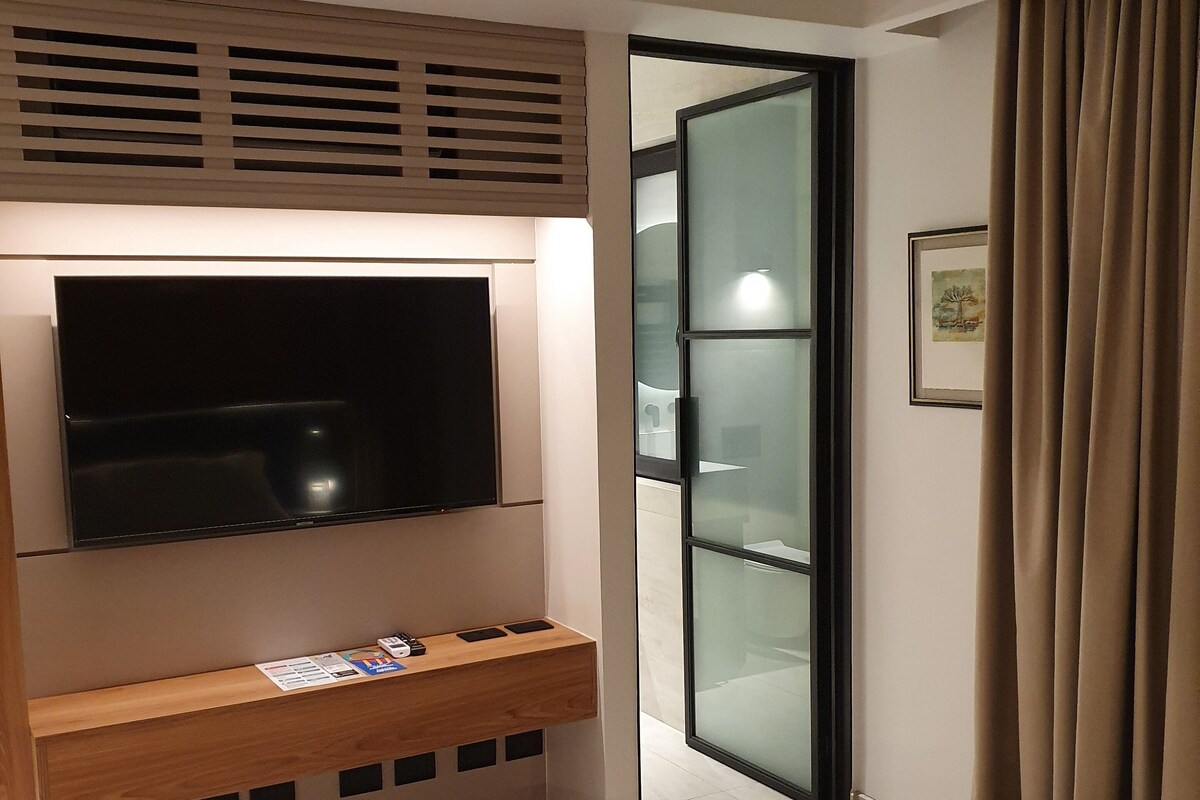 A modern hotel room with a large flat-screen TV mounted on a wooden console, a glass door leading to a bathroom, and a framed picture on the wall.
