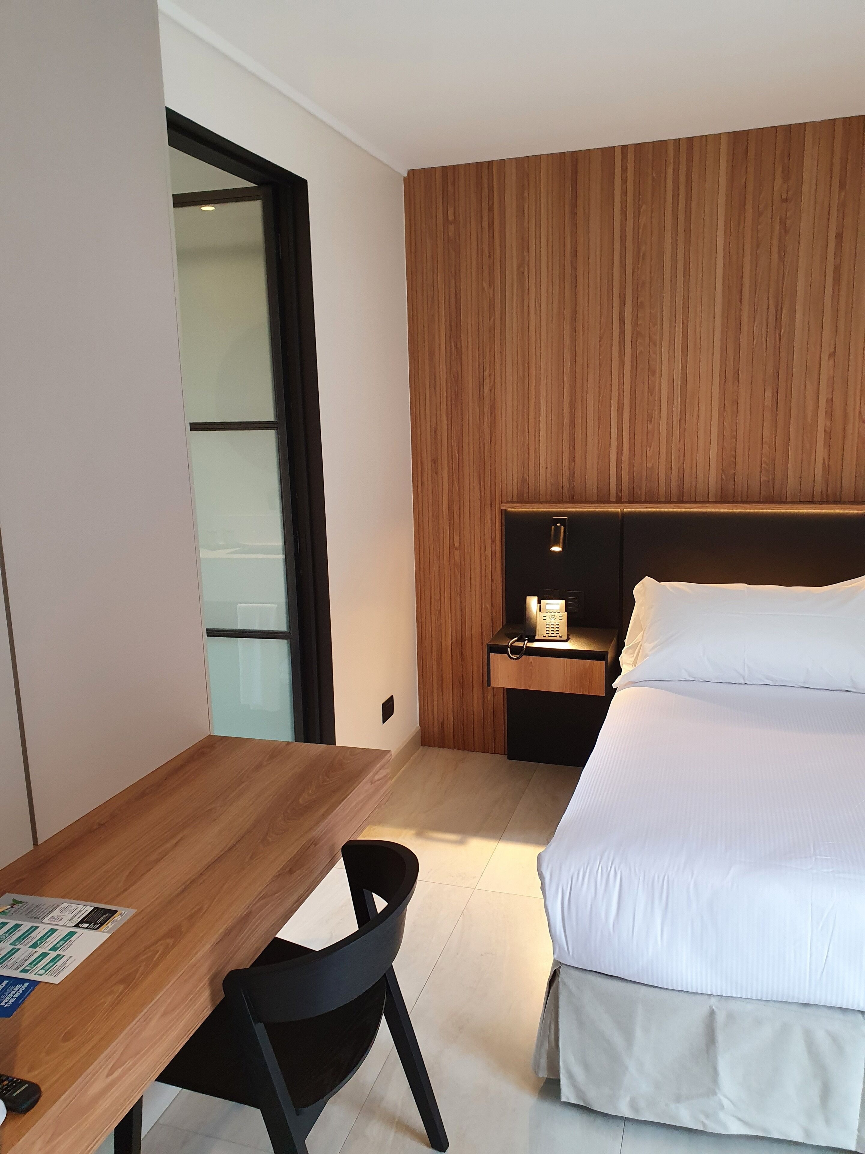 Una habitación de hotel moderna con cabecera de madera, cama con ropa blanca, escritorio con silla y mesita de noche con teléfono.