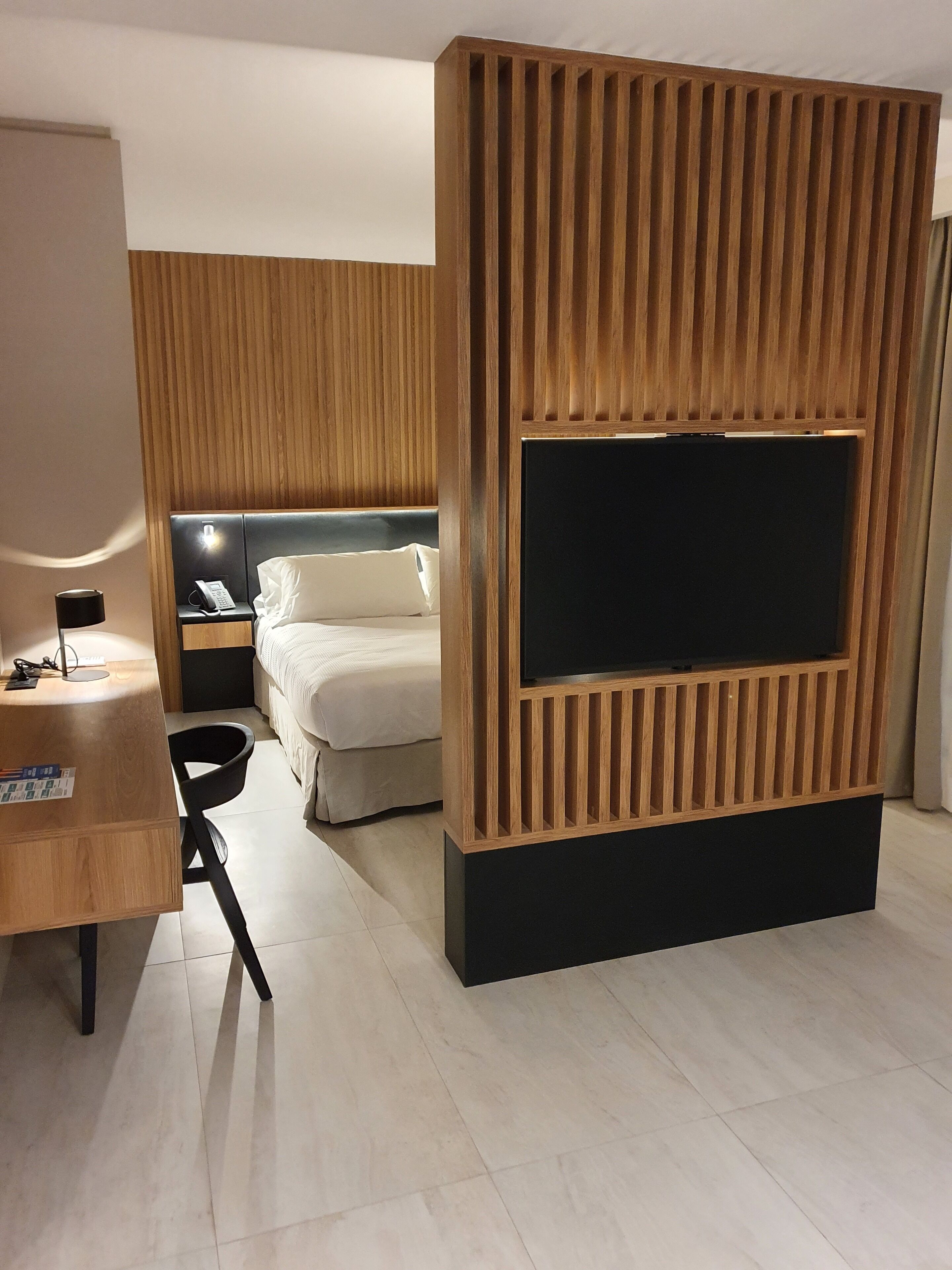 A modern hotel room with a wooden accent wall, a flat-screen TV mounted on a black stand, a bed with white linens, a desk with a chair, and a nightstand with a lamp.