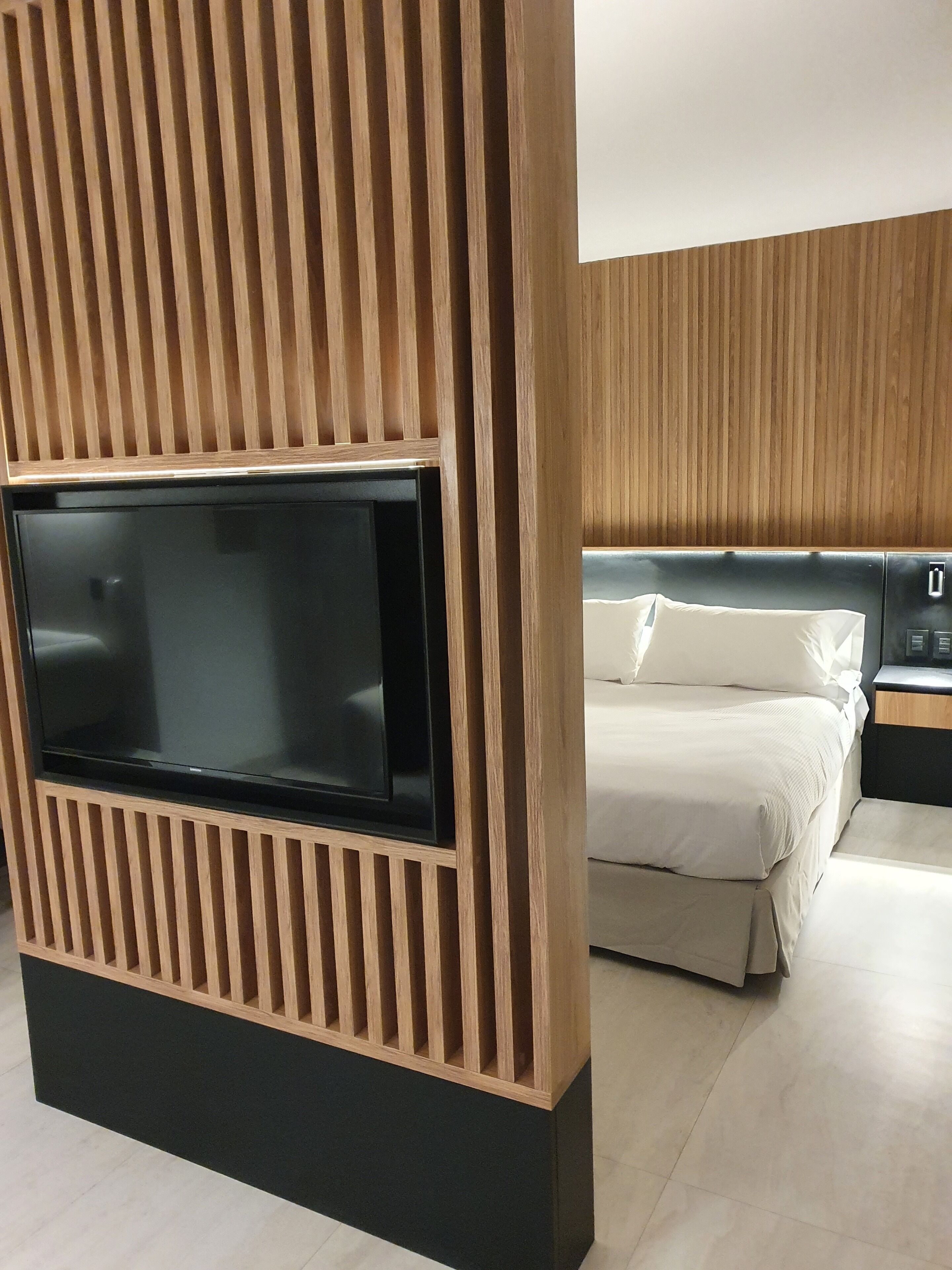 A modern hotel room with a large flat-screen TV mounted on a wooden panel wall, a bed with white bedding, and a nightstand with a lamp.