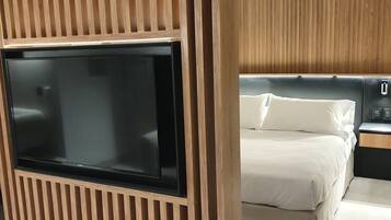 A modern hotel room with a large flat-screen TV mounted on a wooden panel wall, a bed with white bedding, and a nightstand with a lamp.