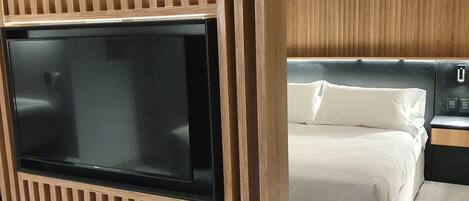 Una habitación de hotel moderna con un televisor de pantalla plana grande montado en una pared de paneles de madera, una cama con ropa blanca y una mesita de noche con una lámpara.