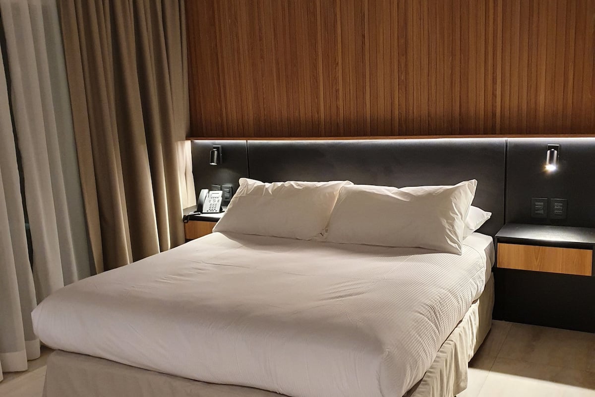 A modern hotel room with a large bed, wooden headboard, and a nightstand with a lamp.