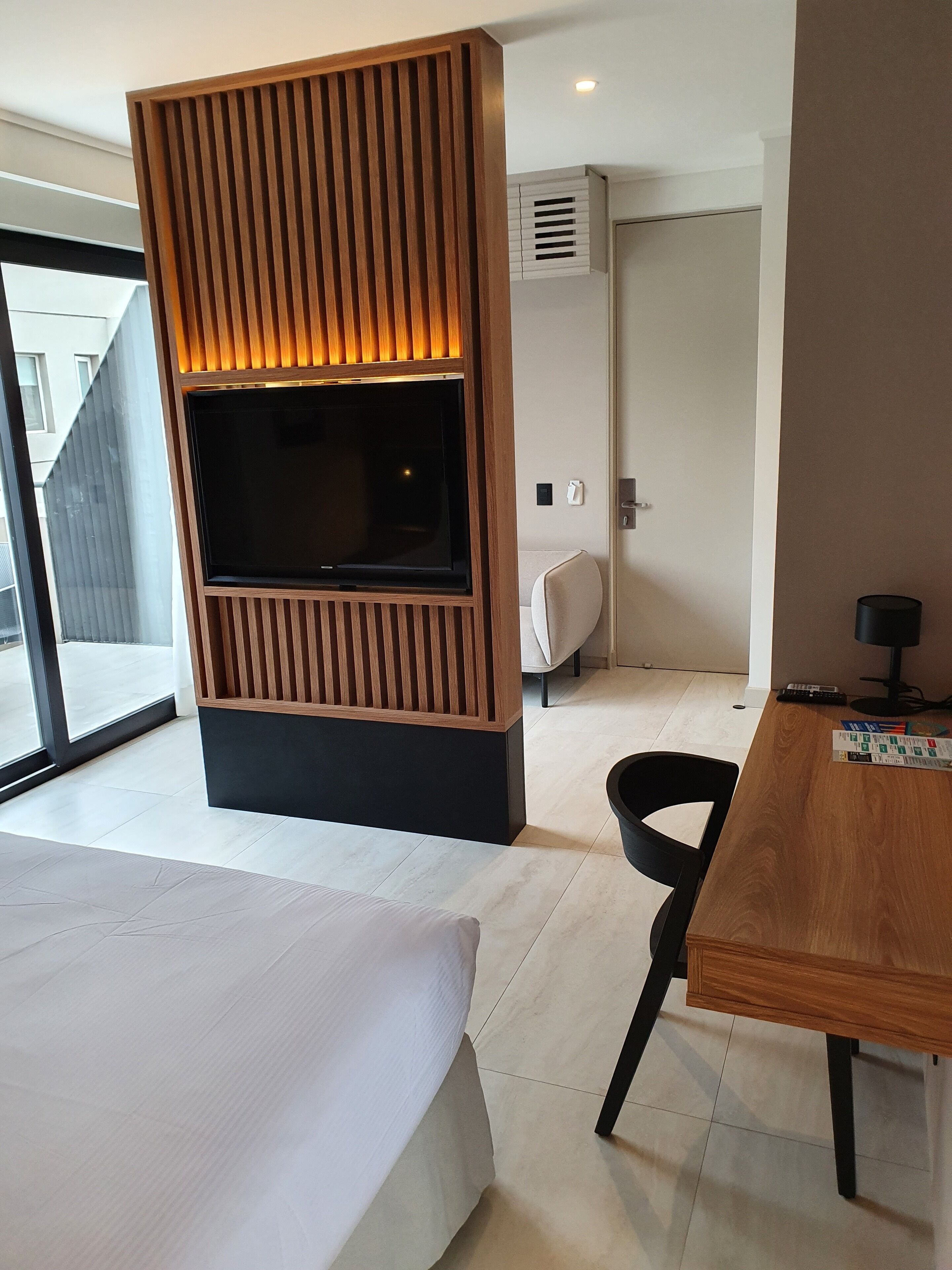 Habitación de hotel moderna con una televisión grande montada en un panel de madera, una cama, un escritorio con una silla y vistas al exterior.