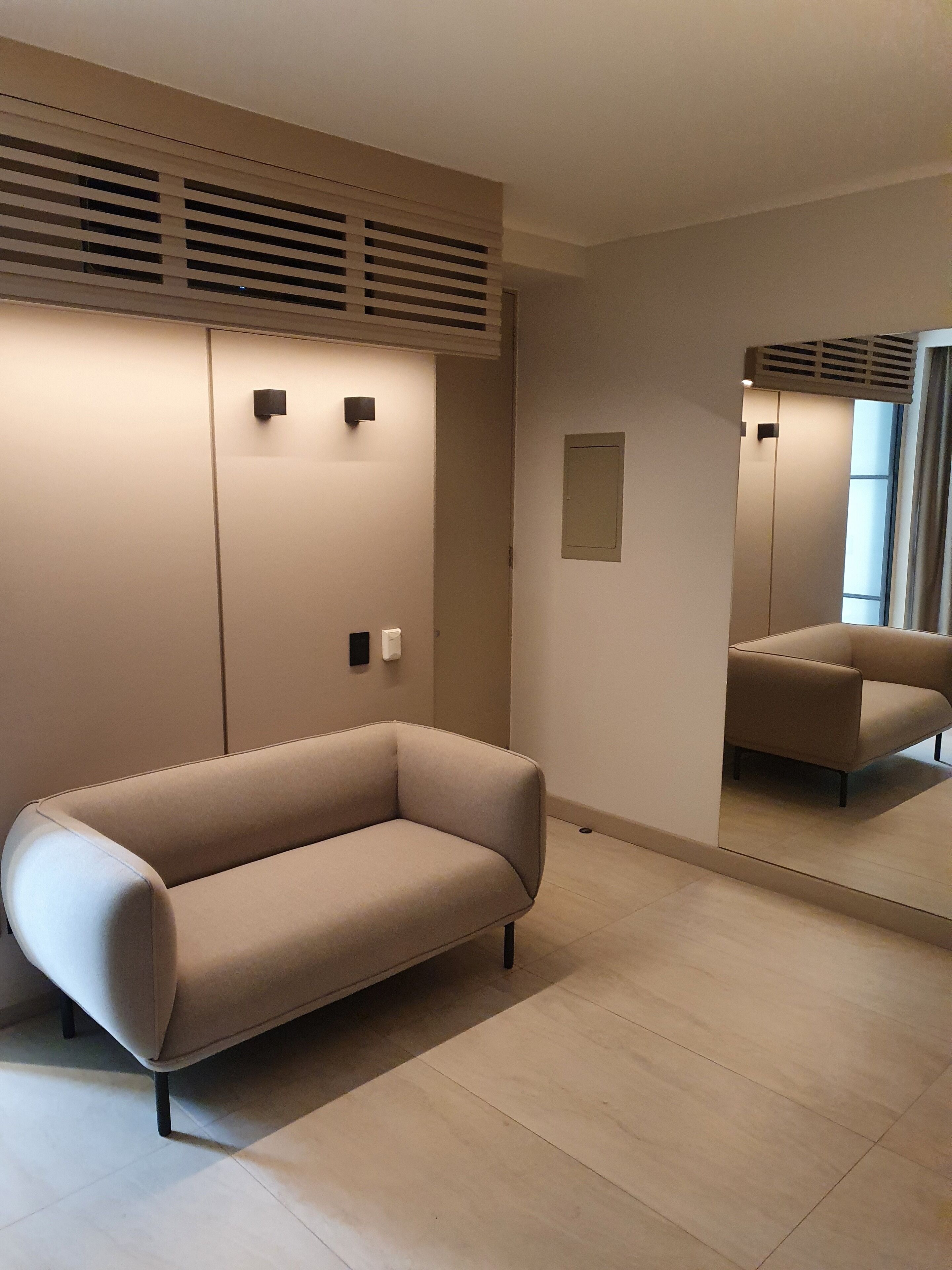 Una habitación moderna con un sofá color beige, un espejo grande y una ventana con persianas.