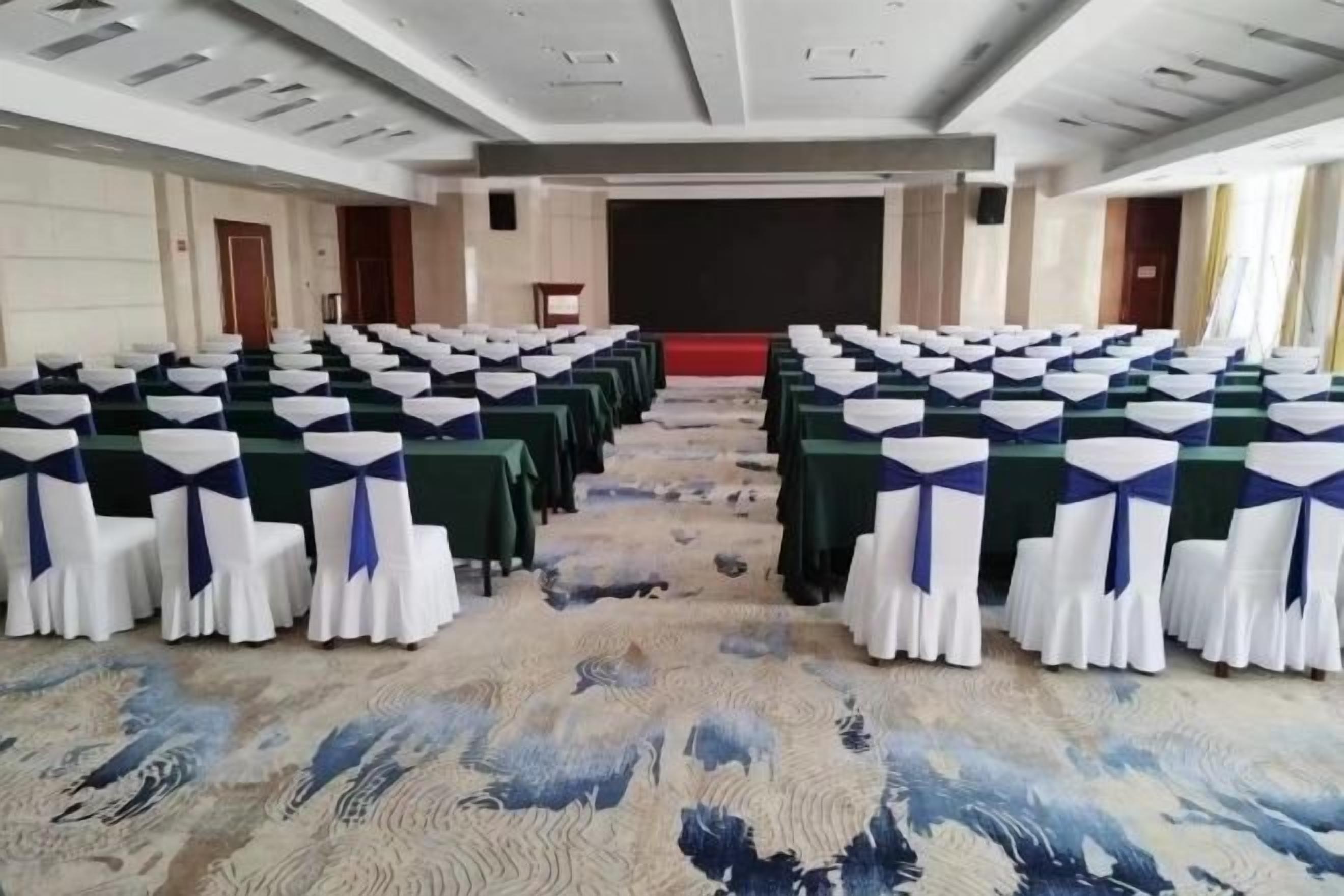A conference room with rows of tables and chairs, a stage, and a carpeted floor.