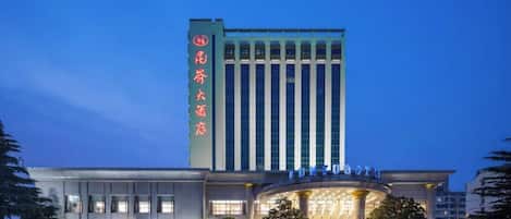 Un hotel con un letrero vertical llamativo, una entrada amplia con techo curvo y un estacionamiento con varios coches.
