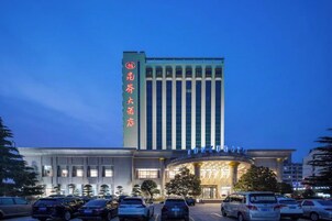 Un hotel con un letrero vertical llamativo, una entrada amplia con techo curvo y un estacionamiento con varios coches.