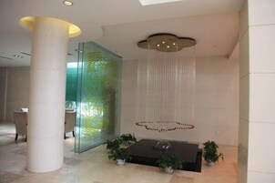 Un vestíbulo moderno con una columna grande, un separador de vidrio con diseño verde, una lámpara colgante y plantas en macetas.