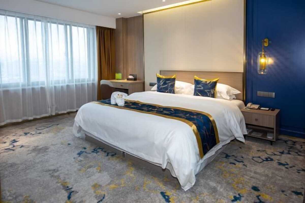 Una habitación de hotel con una cama grande, una mesita de noche con lámpara, y una ventana con cortinas.