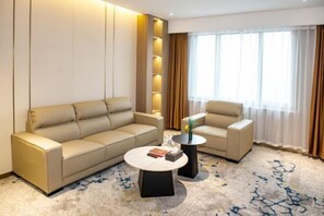 Una habitación de hotel moderna con un sofá color beige, un sillón más pequeño, una mesa de centro redonda y un ventanal con cortinas.