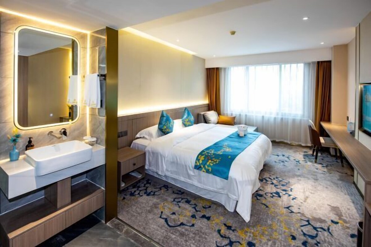 Una habitación de hotel moderna con una cama grande, una mesita de noche, un escritorio, una silla, un espejo amplio, un lavamanos y una ventana con cortinas.