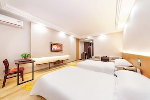 Habitación de hotel con dos camas, un escritorio con una silla, una mesita con una planta, un banco y un cuadro en la pared.