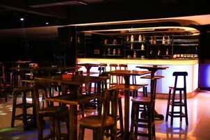 Zona de bar con taburetes y mesas, una barra con botellas y un fondo oscuro.