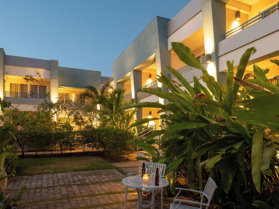 Un patio con un sendero pavimentado, una zona de estar con sillas y una mesa, y un edificio con varios balcones e iluminación.