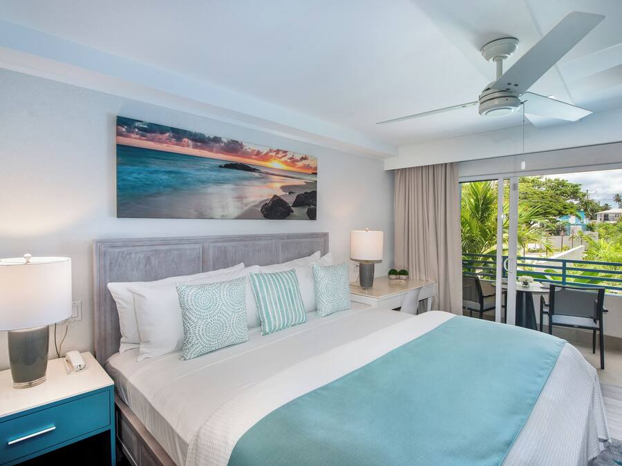 A hotel room with a bed, nightstand, lamp, and a view of a balcony with outdoor furniture.