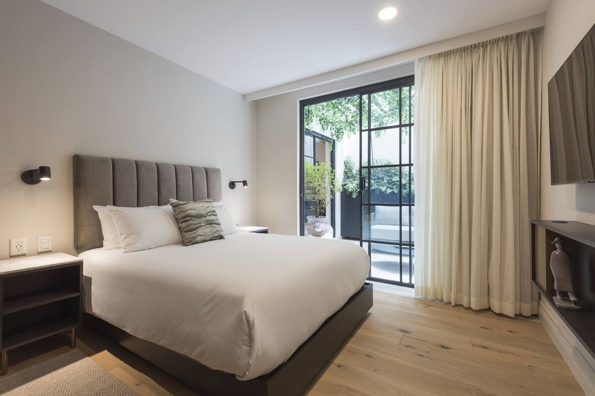 Ein modernes Schlafzimmer mit einem großen Bett, einem Nachttisch, einem am Wand montierten Fernseher und einer Schiebetür zu einem Außenbereich.