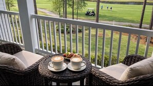 Varanda com mobiliário de vime, uma mesa com duas chávenas de café e croissants, e vista para um campo de golfe com pessoas e um carrinho de golfe.