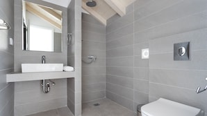 A modern bathroom with a white sink, mirror, and toilet, and a shower area with grey tiles. - Raise Spetses Villa Yasemi (Spetses)