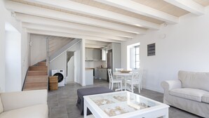 A modern living room with a glass coffee table, a sofa, a dining area with a table and chairs, a kitchen in the background, and a staircase leading to another level.