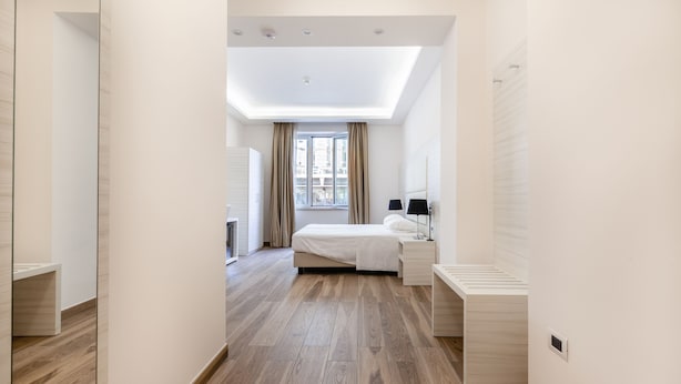 A modern hotel room with a wooden floor, a bed with a white headboard, a bedside table with a lamp, and a window with curtains. - HUB NAPLES (Naples)