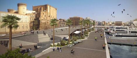 A waterfront promenade with palm trees, a castle-like building, and a marina with boats.