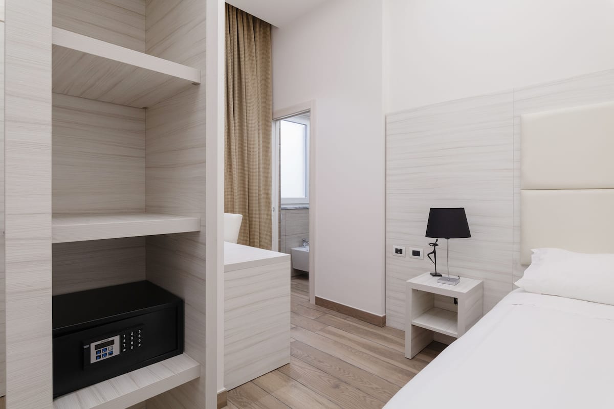 Habitación de hotel con cama, mesita de noche con lámpara, armario y baño visible a través de una puerta abierta.