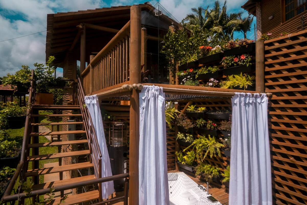 A wooden deck with a staircase leading to a balcony, surrounded by lush greenery and a clear blue sky.