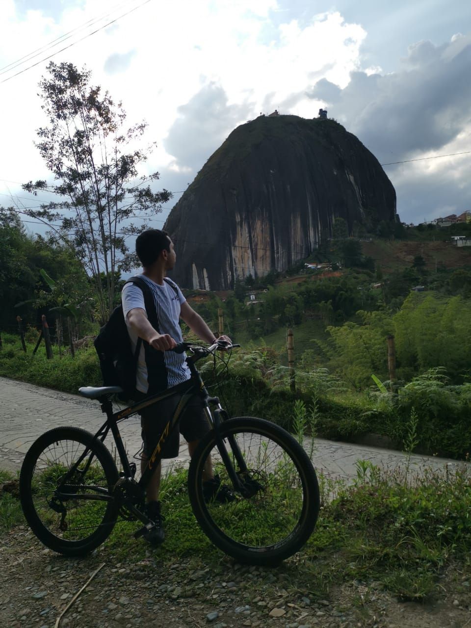 A person on a bicycle with a backpack, a road, and a large rock formation in the background.