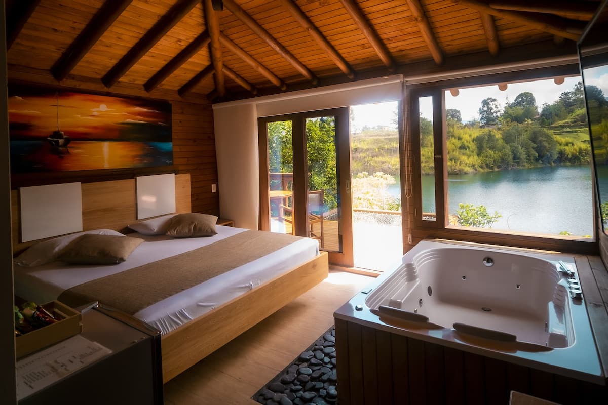A bedroom with a wooden ceiling, a bed, a bathtub, and a view of a lake.