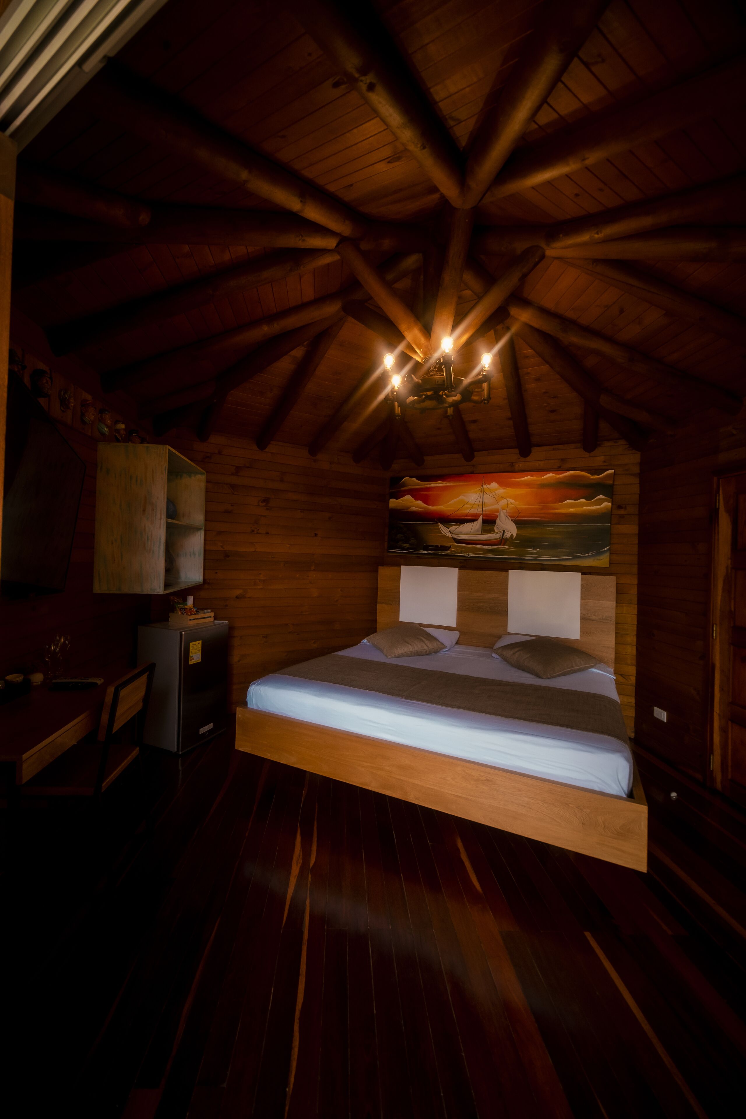 Een slaapkamer in een houten hut, met een bed, een bureau en een schilderij van een ondergaande zon aan de muur.