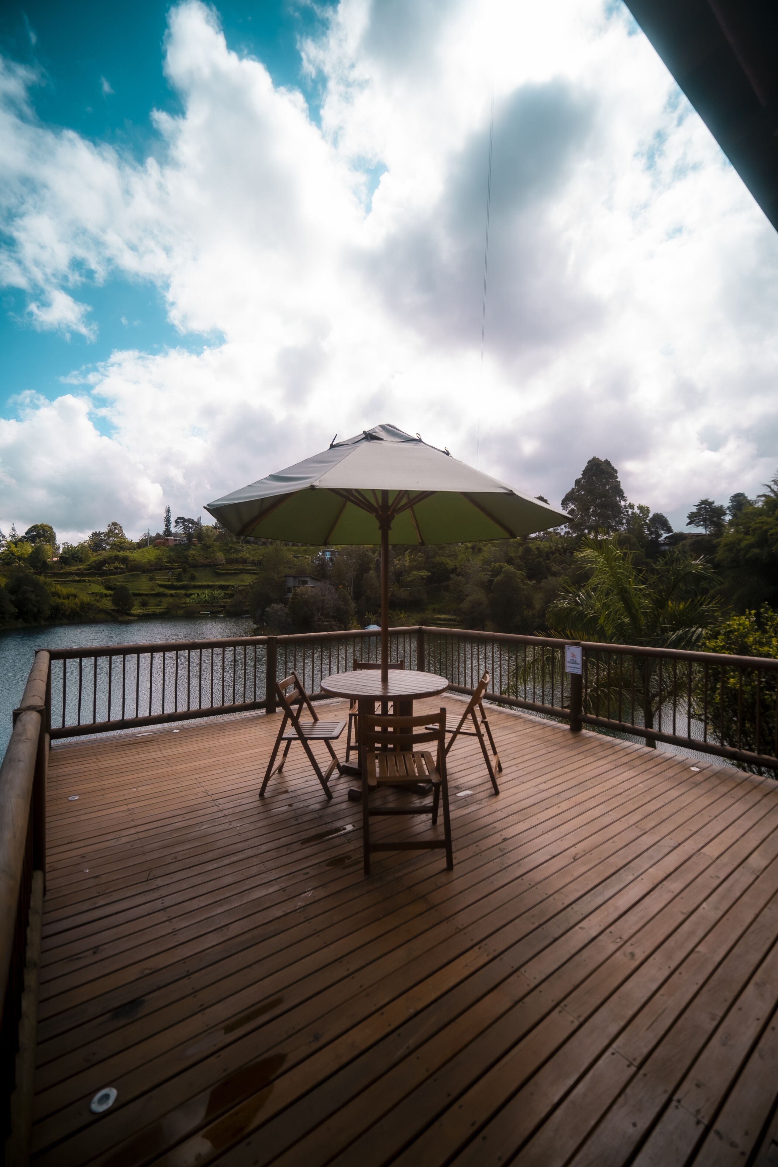 A wooden deck with a table and chairs under a large umbrella, overlooking a body of water and greenery.