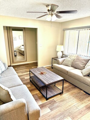 A living room with a sofa, a coffee table, a ceiling fan, and a window with blinds.