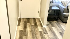 A hallway with a wooden floor leading to a door, a small table, and a gray sofa with a patterned pillow. - Beautiful newly remodeled 3 bed 2 bath foothills home.  No cleaning fees. (Albuquerque)