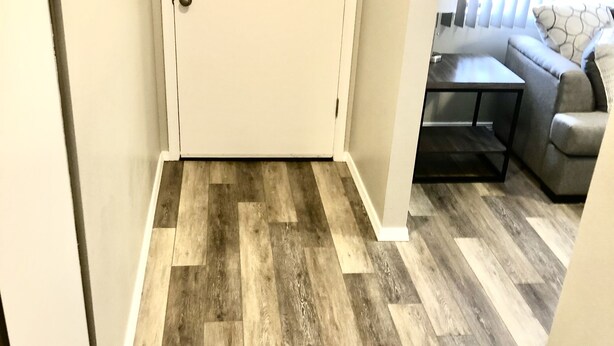 Un couloir doté d’un plancher en bois menant à une porte, à une petite table et à un canapé gris agrémenté d’un coussin à motifs.