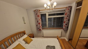 A neatly made bed with a white duvet, two pillows, and a rolled towel. A wooden headboard, a bedside table, and a mirror are also visible.