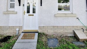 Un edificio de dos pisos con fachada blanca, techo negro y un pequeño porche con techo triangular. En cada piso hay dos ventanas y una puerta con manija de latón.