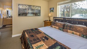 A bedroom with a large bed, a nightstand, a lamp, a painting, and a window with a view of trees and a car.