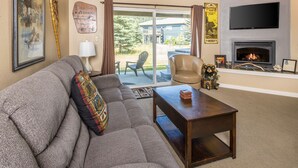 A living room with a grey sofa, a wooden coffee table, a fireplace, a television mounted above it, and a sliding glass door leading to an outdoor area.