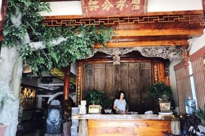 A reception desk with a person standing behind it, a wooden door with traditional carvings, and a hanging plant above.